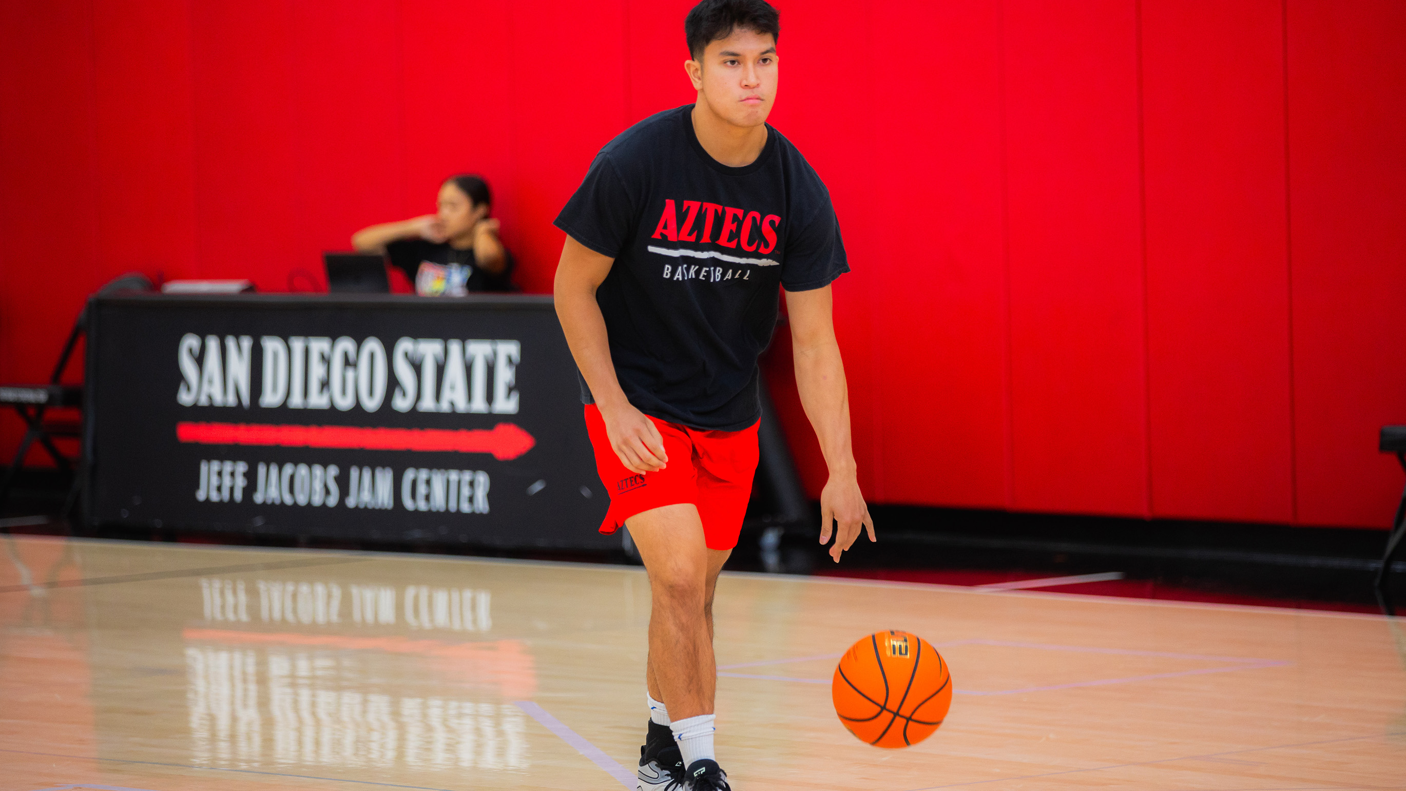 A young man in a black Aztecs Basketball t-shirt and red shorts softly dribbles a basketball on a court with a table identified as Jeff Jacobs Jam Center behind him.