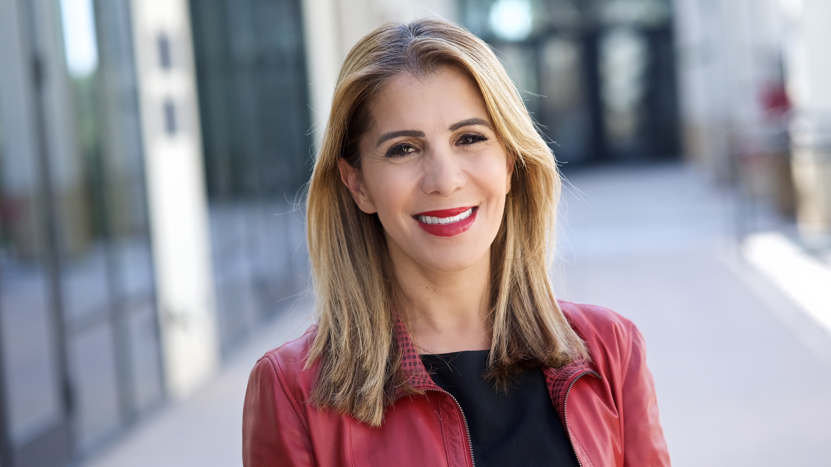 A woman with shoulder-length blond hair, wearing a cranberry jacket over a black top, is smiling with blurred glass windows and doors in nthe background.