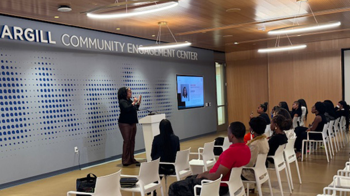 A classroom with about a dozen students seated in white chairs, facijng a speaker at a white podium in front of a sign reading Community Engagement Center.