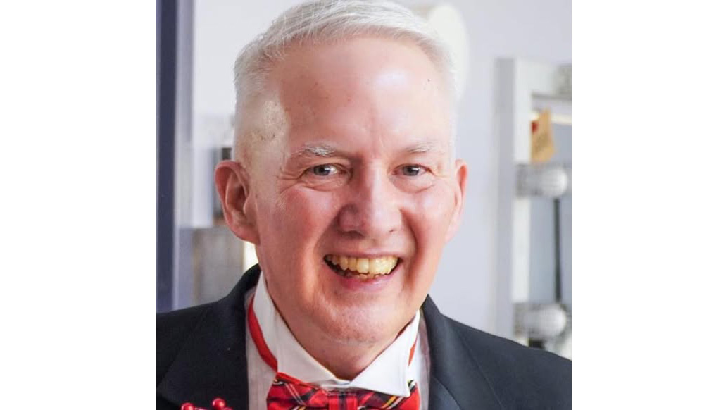 A portrait of a man smiling broadly, wearing a dark coat and red plaid bowtie.