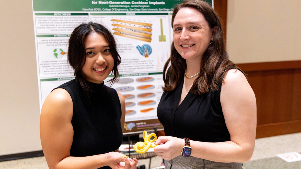 Two young women wearing black dresses pose in front of a science poster titled Next-Generation Cochlear Implants. Each is holding a piece of a model of the implants.