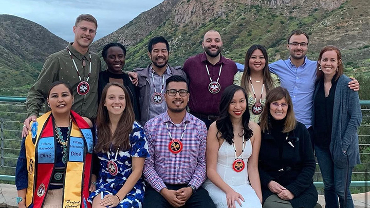 Twelve men and women in two rows with the front row seated on a low barrier with a fence overlooking a long, deep canyon. Many are wearing tribal necklaces.