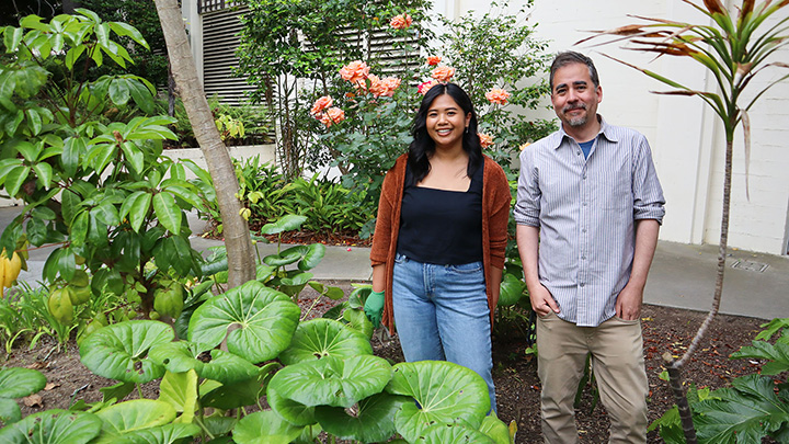 A woman wearing a black top and brown jacket and a man in a gray shirt are standing in the dirt of a garden with large green plants. 