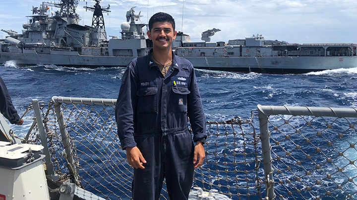 A man in a flight suit stands on the deck of a ship in the ocean alongside a  gray warship.