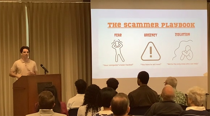 A young man in a golden polo shirt stands at a podium addressing a seated audience of a dozen or more people next to a screen displaying the words The Scammer Playbook Fear Urgency Isolation.
