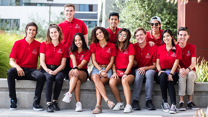 Eleven college-age men and women, all wearing red polo shorts, pose in two rows with the front row seated on a concrete bench.