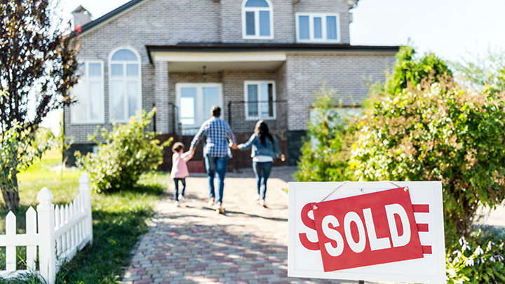 A family of three is seen from the rear as they walk up a cobblestone pathway to a two-story house. A sold sign stands in the foreground.