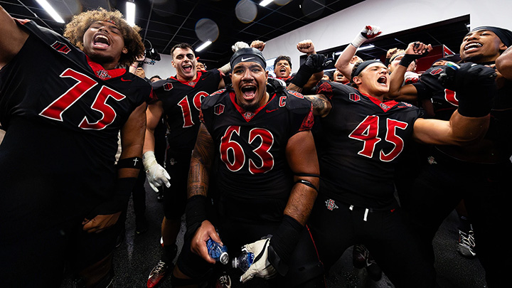 More than a dozen members of a college football team wearing red and black jerseys gesture and yell triumphantly in an indoor room.