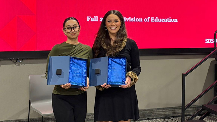 Two women holding awards cases stand side by side in front of a screen displaying a partially obscured message that reads Fall 2025 Division of Education.