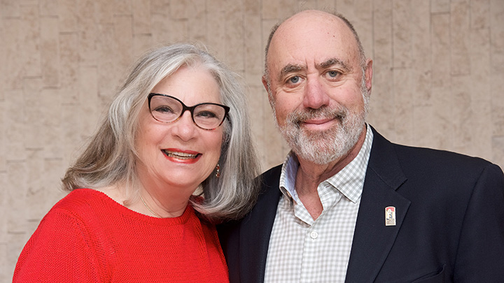 A portrait of a couple. The woman on the left is wearing a red top and glasses. The man has a blazer, a checked dress shirt and a lapel pin.