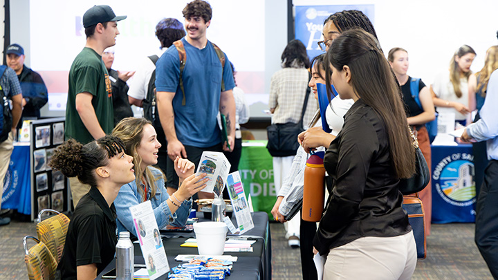 Two women seated behind a table covered with pamphlets and converse with three young women as others mill about in the background with additional tables.