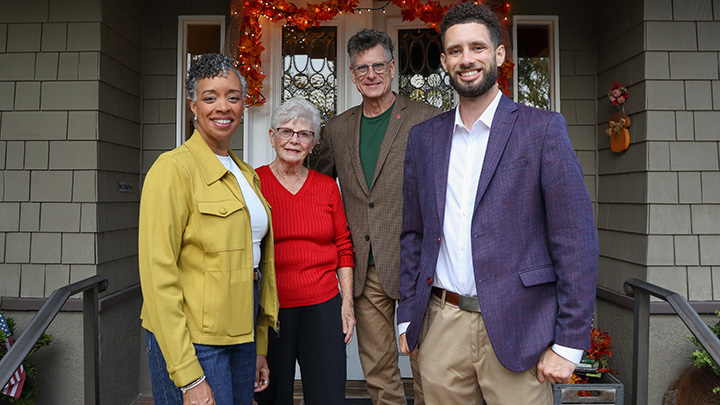 Four people ranging in age from young man to senior woman are standing on the front porch steps of a home with a lighted holiday garland over the door.