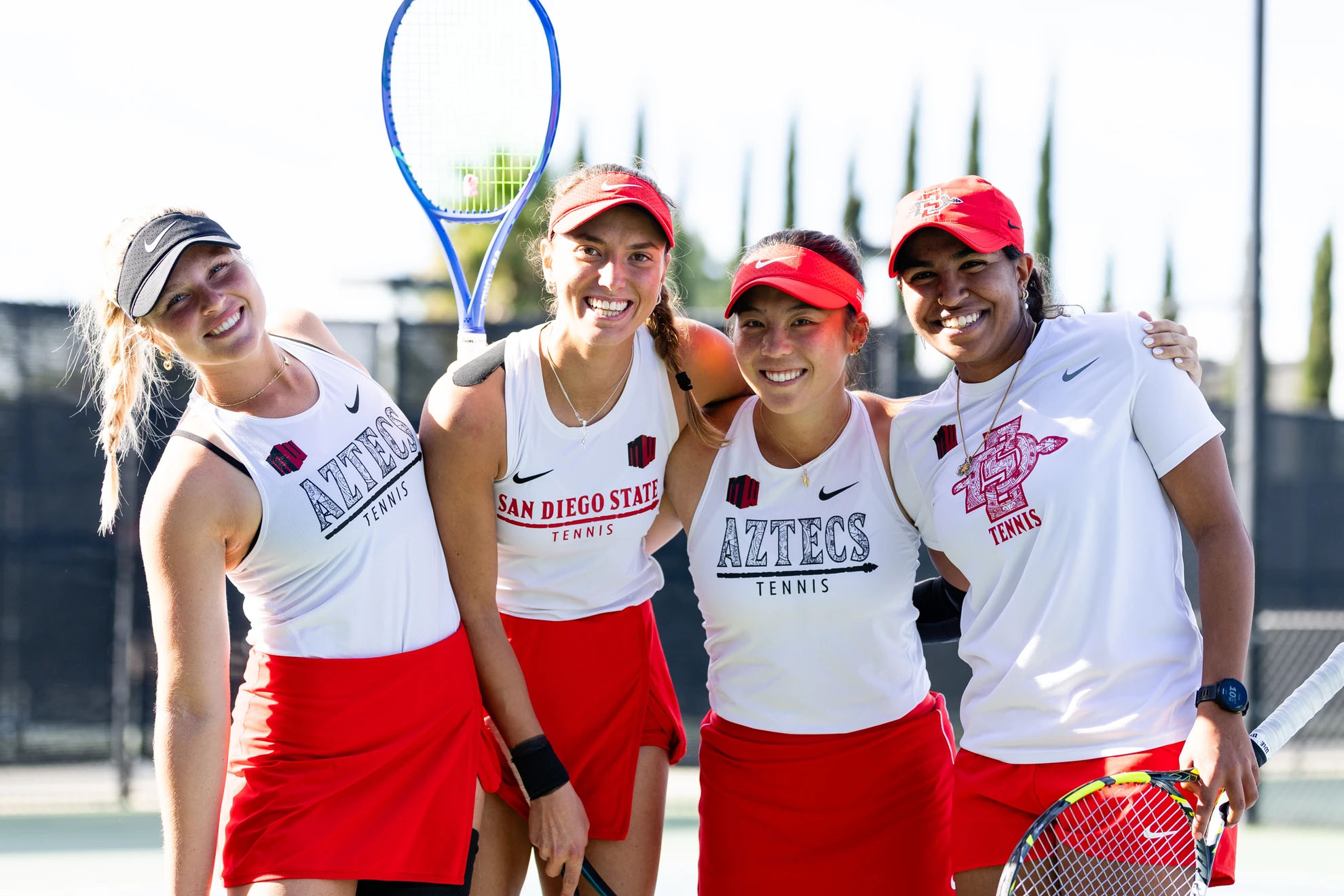 Four women in white tank tops identifying themselves as SDSU tennis team members, and holding tennis racquets, smile with their arms around one another.