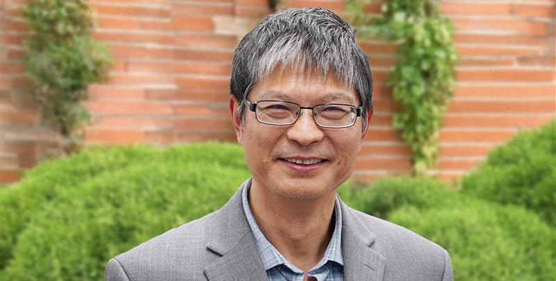 A close-up portrait of a man wearing a gray jacket, with dome-shaped topiary and a brick wall in the background.