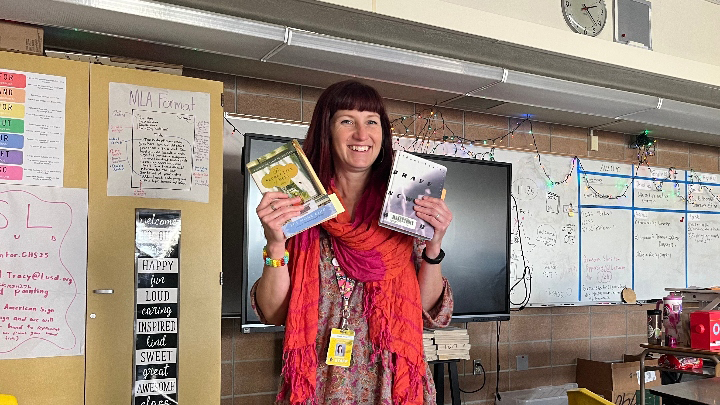 A woman wearing a bright red decorative scarf stands in the front of a classroom holding up a book in each hand.