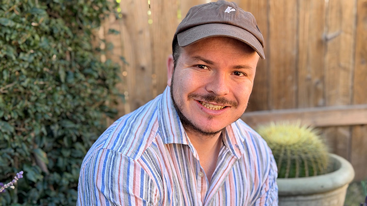 A close-up portrait of a man in a striped, open-collar whirt with a brown cap. Behind him are a large cactus in a planter and a wooden plank fence.