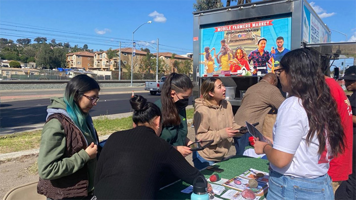 Seven people are gathered around a table in a parking lot along a city street with a truck behind them that reads Mobile Farmers Market on the back panel.