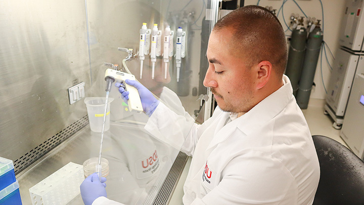 A man in a white lab coat holds a medical instrument with a long plastic syringe, with other assorted medical equipment at his station.