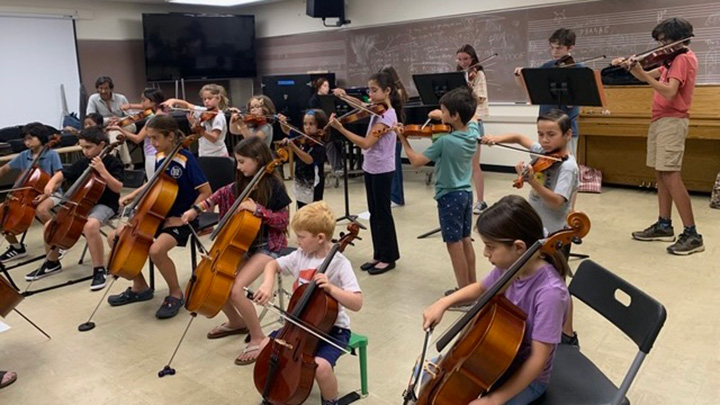 At least 16 young students, both seated and standing, are in a music class playing string, wind and brass instruments.
