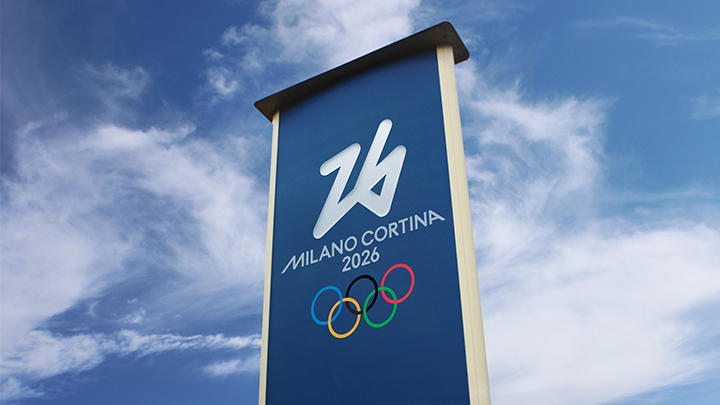 A towering blue monument sign that reads Milano Cortina 2026, featuring the Olympic rings logo, with white clouds and a blue sky behind it.