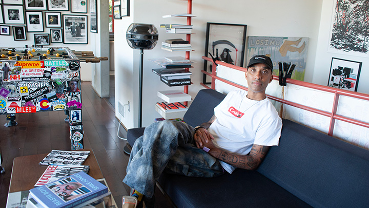 A man wearing a white t-shirt with a Canon logo and a black baseball cap sits on a sofa with his hands folded on his lap, near a foosball table plastered with stickers on the side.