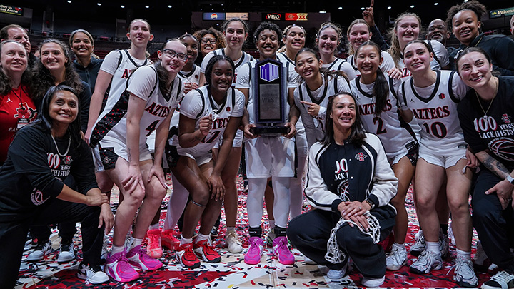 A team of women in white Aztecs basketball jerseys gather around a player holding a trophy in an arena.