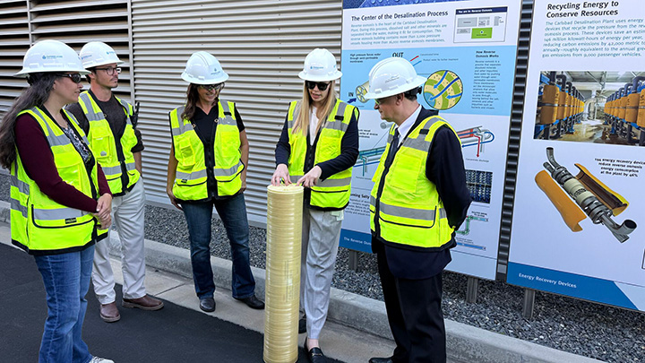 Five people in reflective safety vests and white hardhats stand around a short cylinder while one of the women is explaining something.