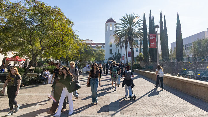 Students are strolling along a long campus pathway with a tower in the deep background and trees and banners along the sides.
