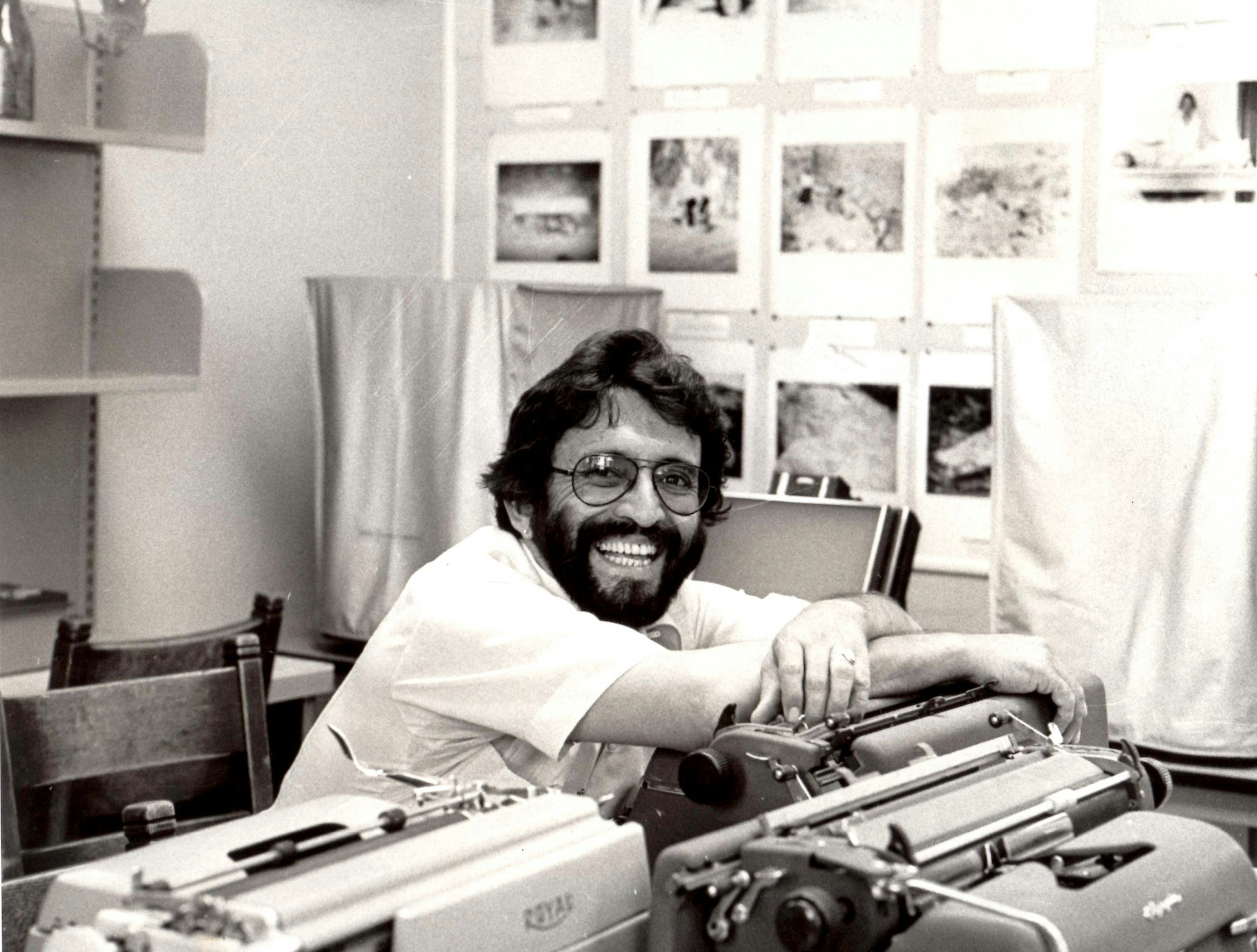 A smiling man sits at a table with his hands crossed over the top of a typewriter.