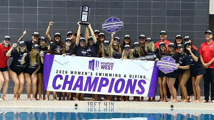 More than two dozen young women stand in a line at the end of a swimming pool with signs, caps and a trophy celebrating a conference championship.and 