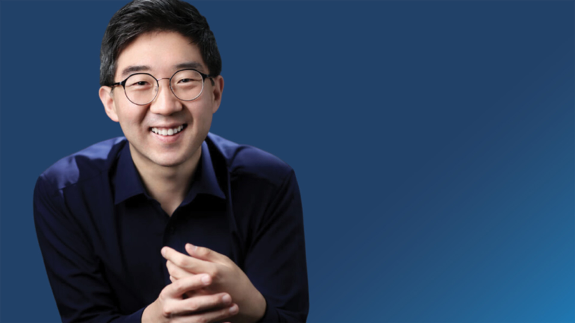 A professional photograph of a broadly smiling man from the elbows up, with short black hair, deep blue shirt and round glasses frames.