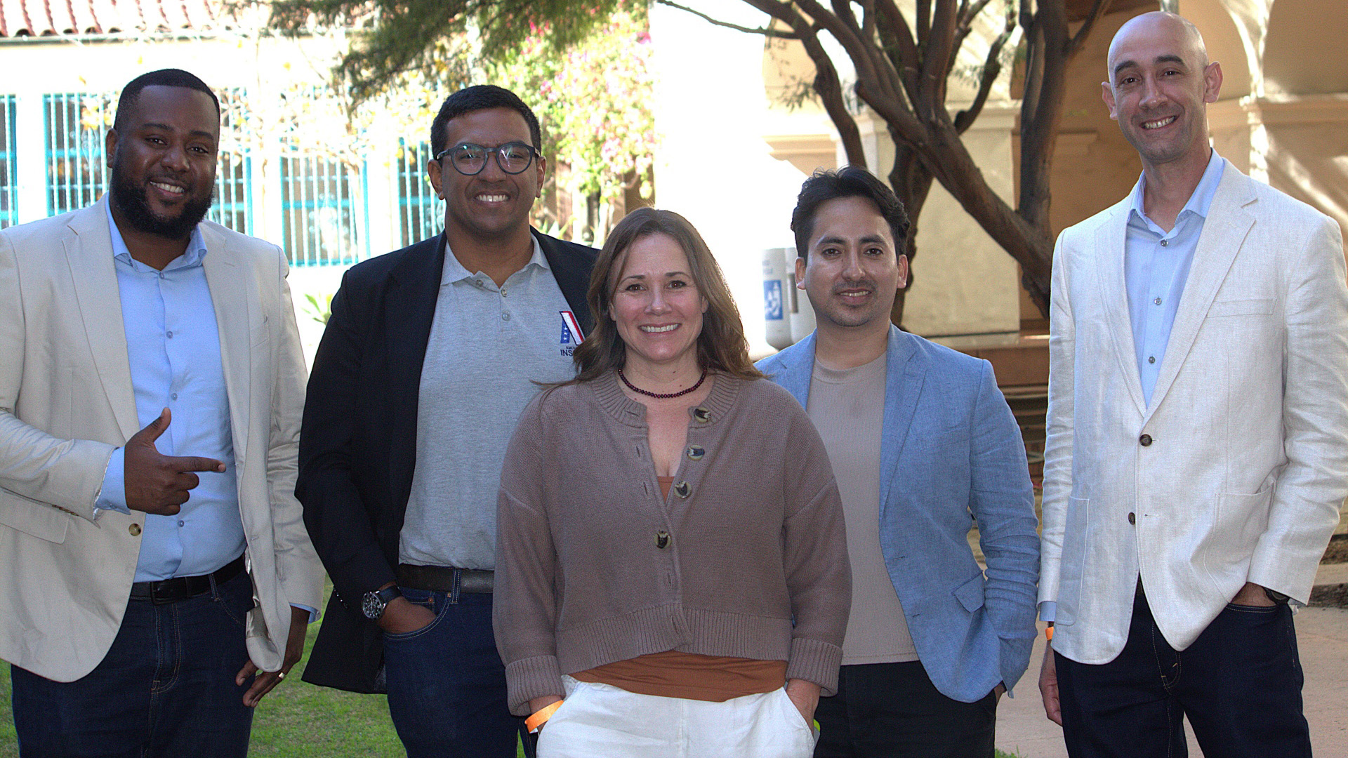 Four men and a woman stand side by side outdoors; the man on the left is pointing playfully at the man to his left.