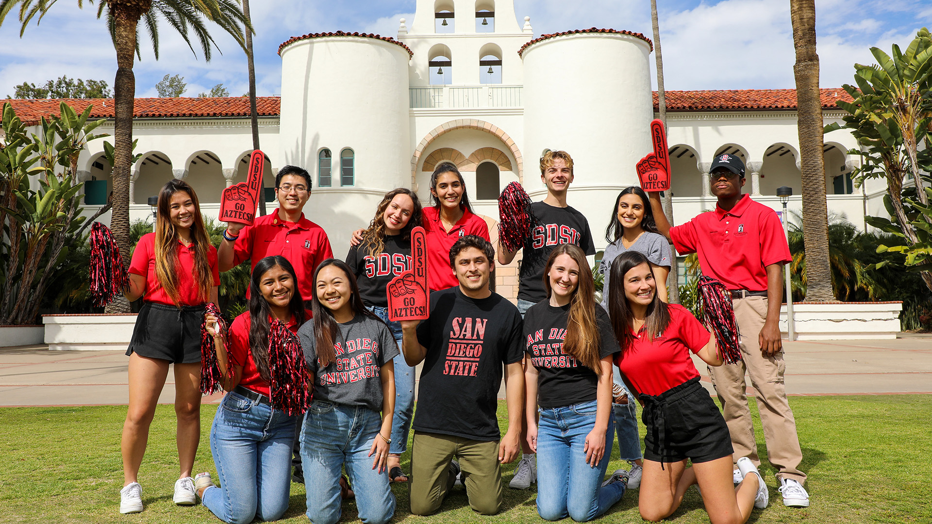 Twelve male and female students with San Diego State University clothing and foam fingers pose in two rows on the lawn in front of a Spanish Revival style building.