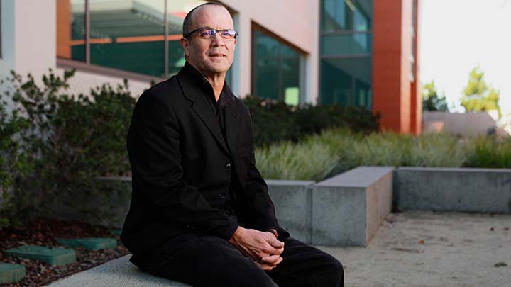 A man in a black suit is sitting on a concrete bench outside a building. His hands are clasped between his legs, and there is some drought-tolerant landscaping in the background next to the building.